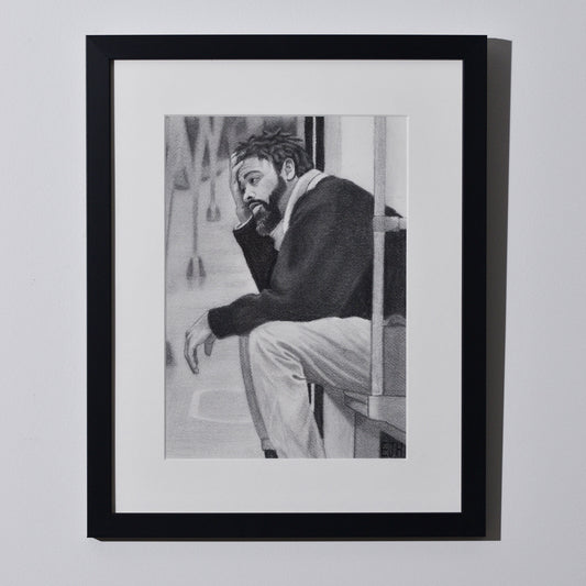 'Lost In My Head' framed portrait drawing of a man on metro seat hanging on a neutral wall in the artist's studio.