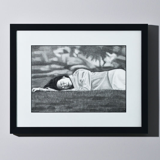 'Passing Of Time' framed portrait drawing of a woman lying on grass hanging on a neutral wall in the artist's studio.
