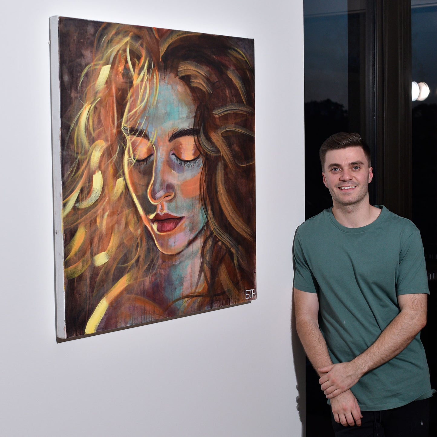 Artist standing next to 'Sunrise' acrylic painting of a woman hanging on wall in studio.