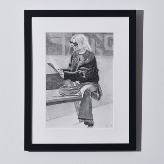 'The Pages We Choose To Fill' framed portrait drawing of a woman reading book hanging on a neutral wall in the artist's studio.