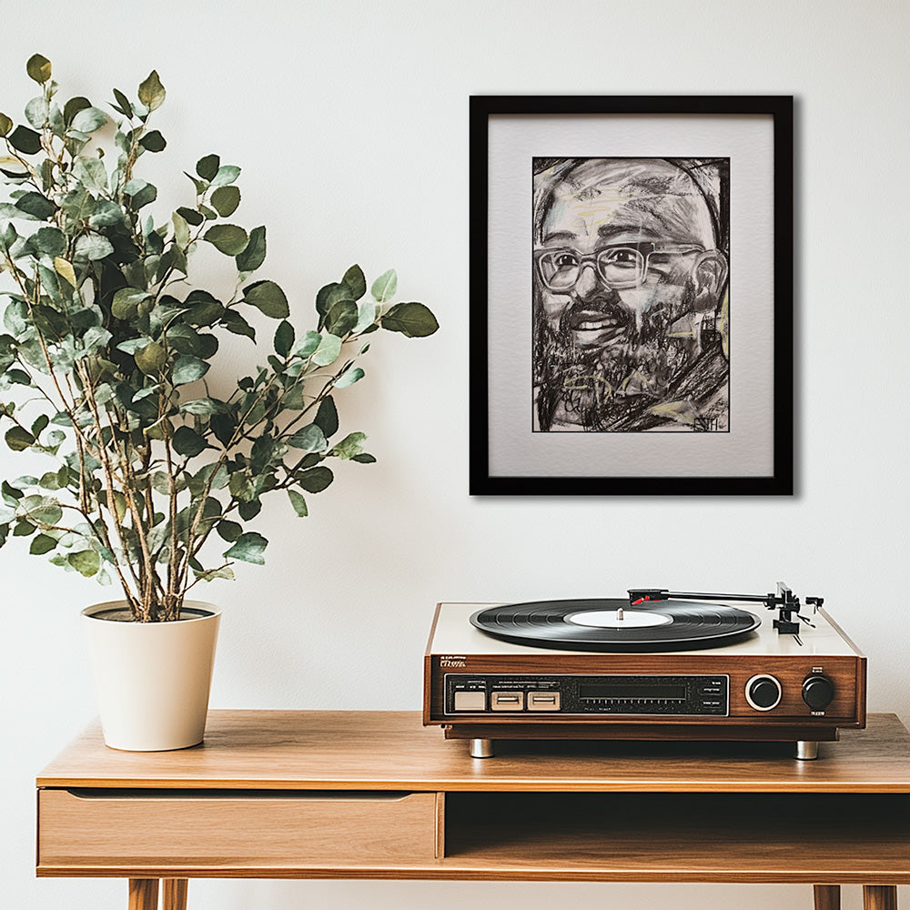 'Amaze' framed portrait drawing hanging above a wooden console table with a turntable and potted plant against a white wall.