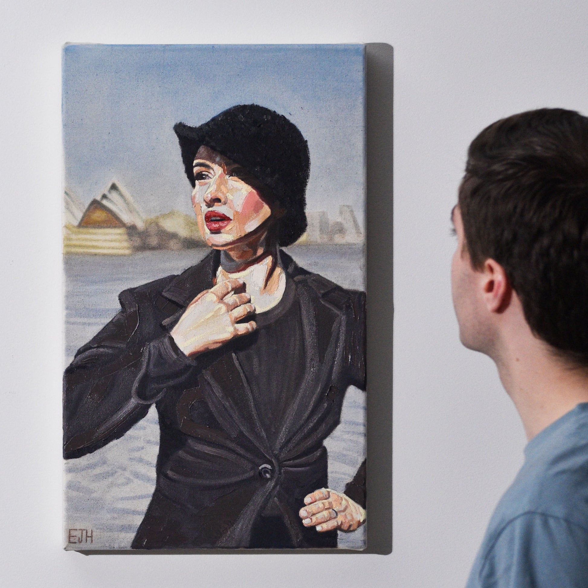 Artist standing next to 'Harbour View' oil painting of a woman with a black blazer and hat standing in front of Sydney Opera House.