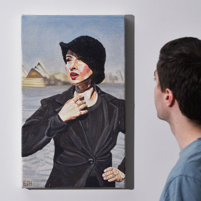 Artist standing next to 'Harbour View' oil painting of a woman with a black blazer and hat standing in front of Sydney Opera House.