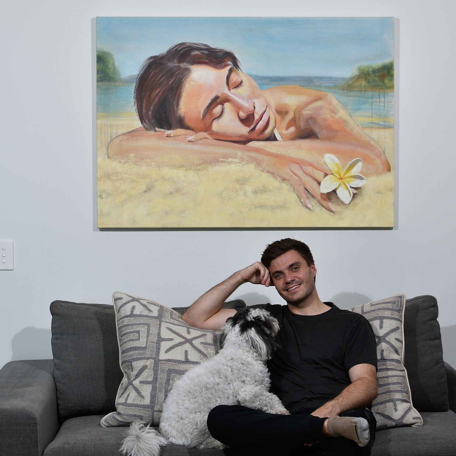 Artist seated next to 'Noon' oil painting of woman holding a frangipani laying on the sand.