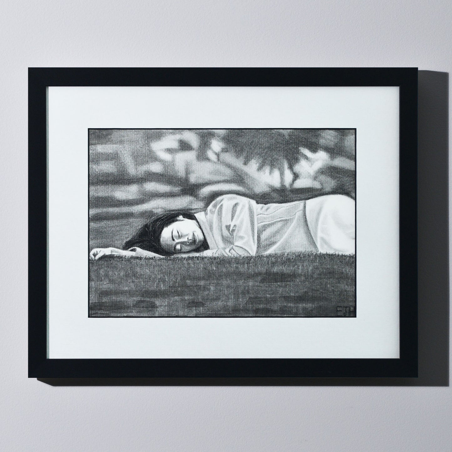 'Passing Of Time' framed portrait drawing of a woman lying on grass hanging on a neutral wall in the artist's studio.