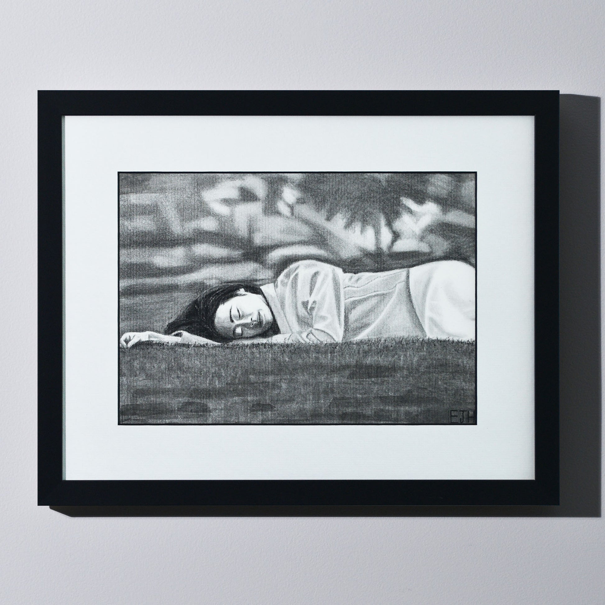 'Passing Of Time' framed portrait drawing of a woman lying on grass hanging on a neutral wall in the artist's studio.