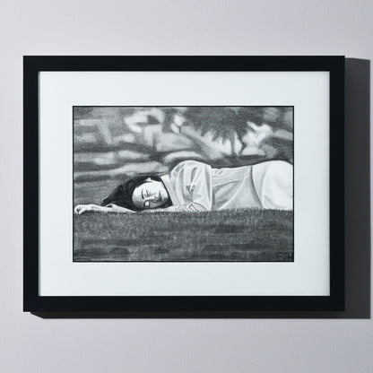 'Passing Of Time' framed portrait drawing of a woman lying on grass hanging on a neutral wall in the artist's studio.