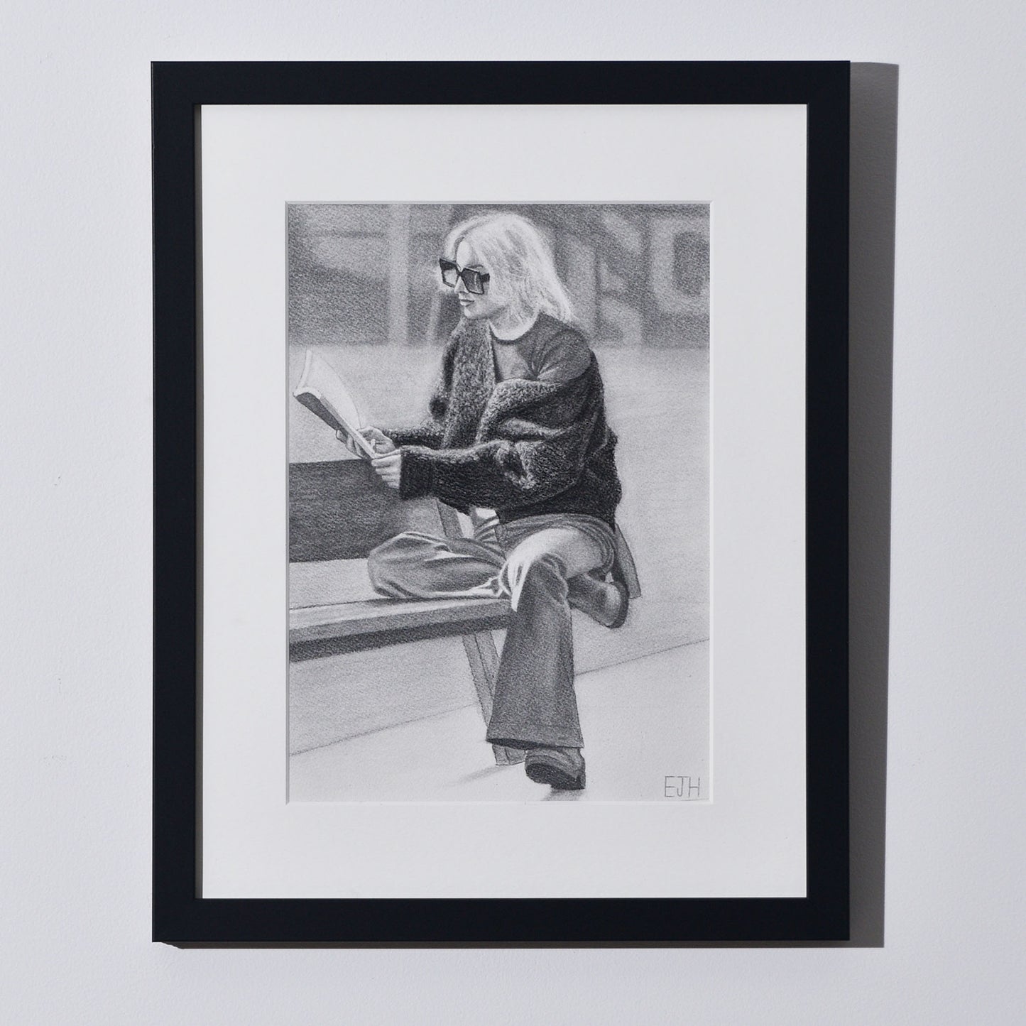 'The Pages We Choose To Fill' framed portrait drawing of a woman reading book hanging on a neutral wall in the artist's studio.