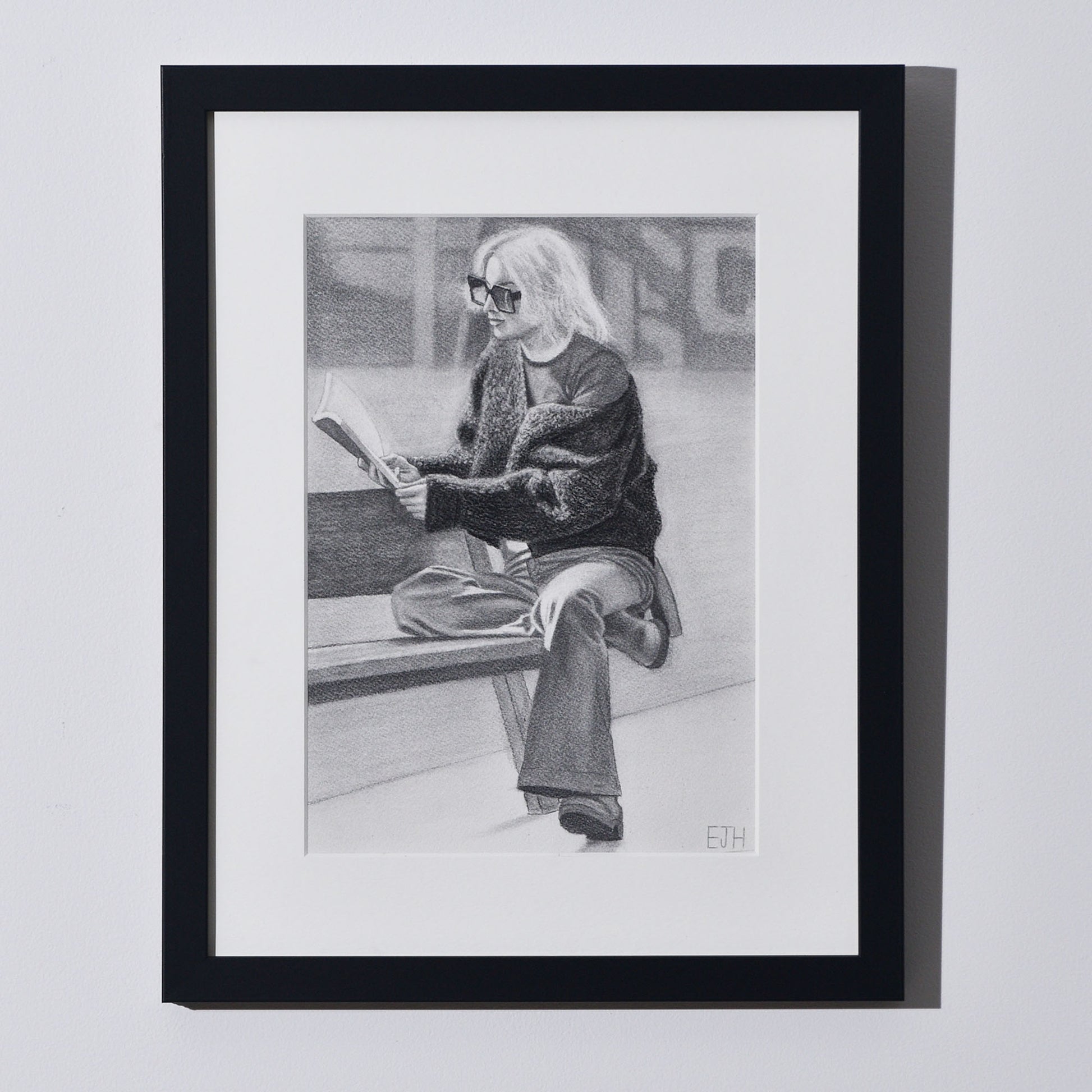 'The Pages We Choose To Fill' framed portrait drawing of a woman reading book hanging on a neutral wall in the artist's studio.