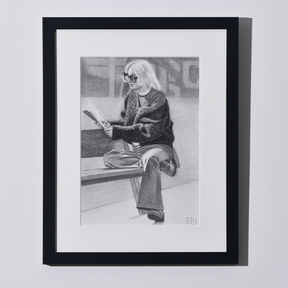 'The Pages We Choose To Fill' framed portrait drawing of a woman reading book hanging on a neutral wall in the artist's studio.