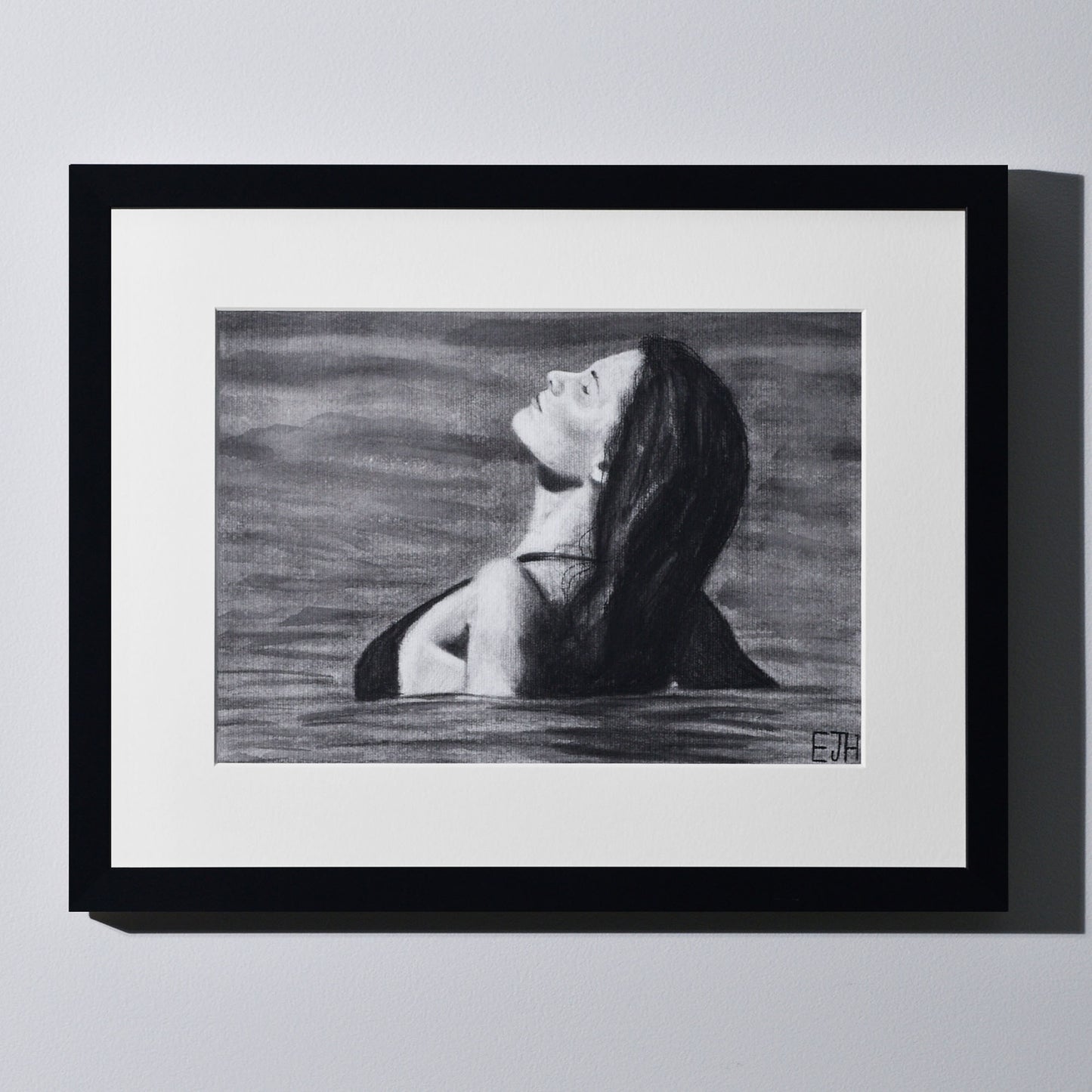 'The Sunbather' framed portrait drawing of a woman in the ocean hanging on a neutral wall in the artist's studio.